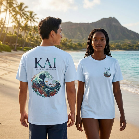 Two models standing on a beach wearing matching light blue t-shirts. The model on the left shows the back of the shirt with a large graphic of a wave and the text 'KAI', while the model on the right shows the front of the shirt with a small chest graphic.