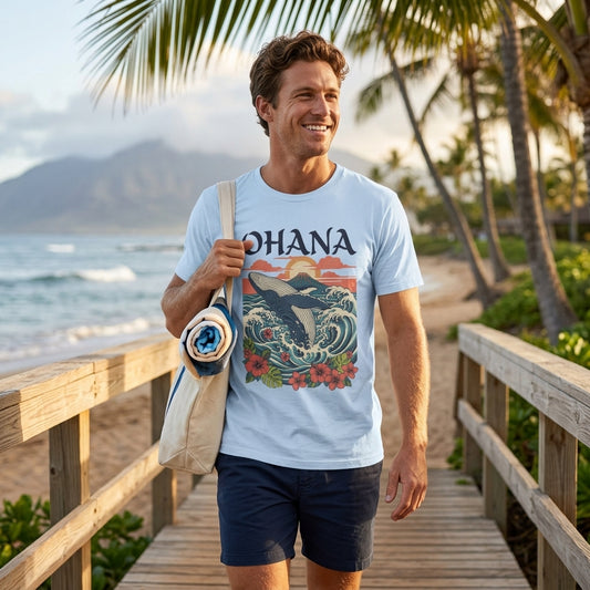 A man walking on a wooden boardwalk by the beach wearing a light blue graphic t-shirt with 'OHANA' and a wave design, navy shorts, and carrying a cream tote bag.