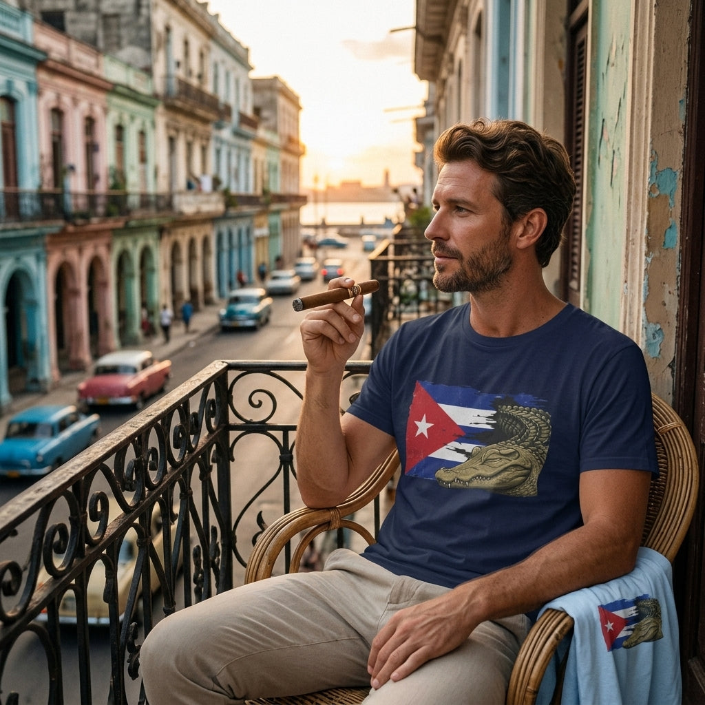 A man sits on a wicker chair on a balcony overlooking a street lined with colorful vintage buildings. He is wearing a navy blue t-shirt with a graphic print of the Cuban flag and a large eagle, with a light blue towel draped over his arm.
