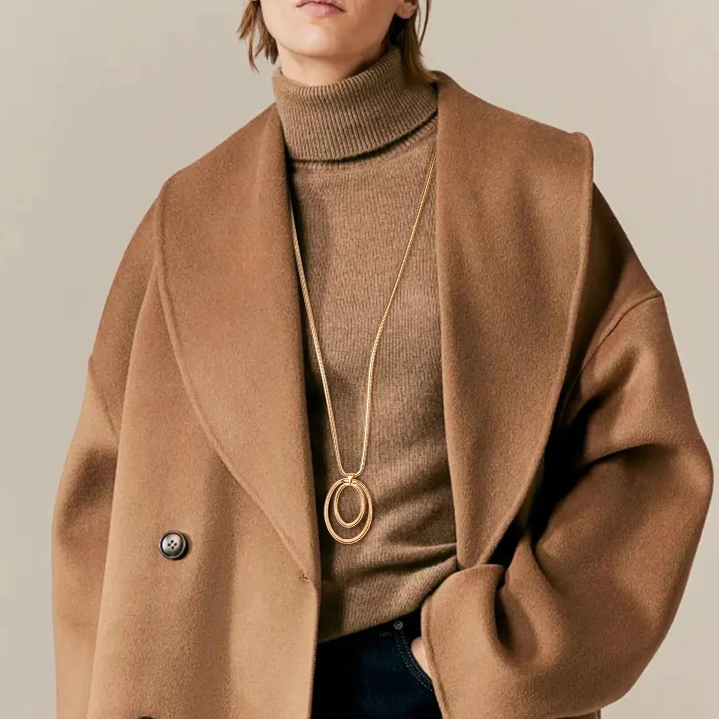 Person wearing a brown coat and turtleneck with a gold necklace on a neutral background