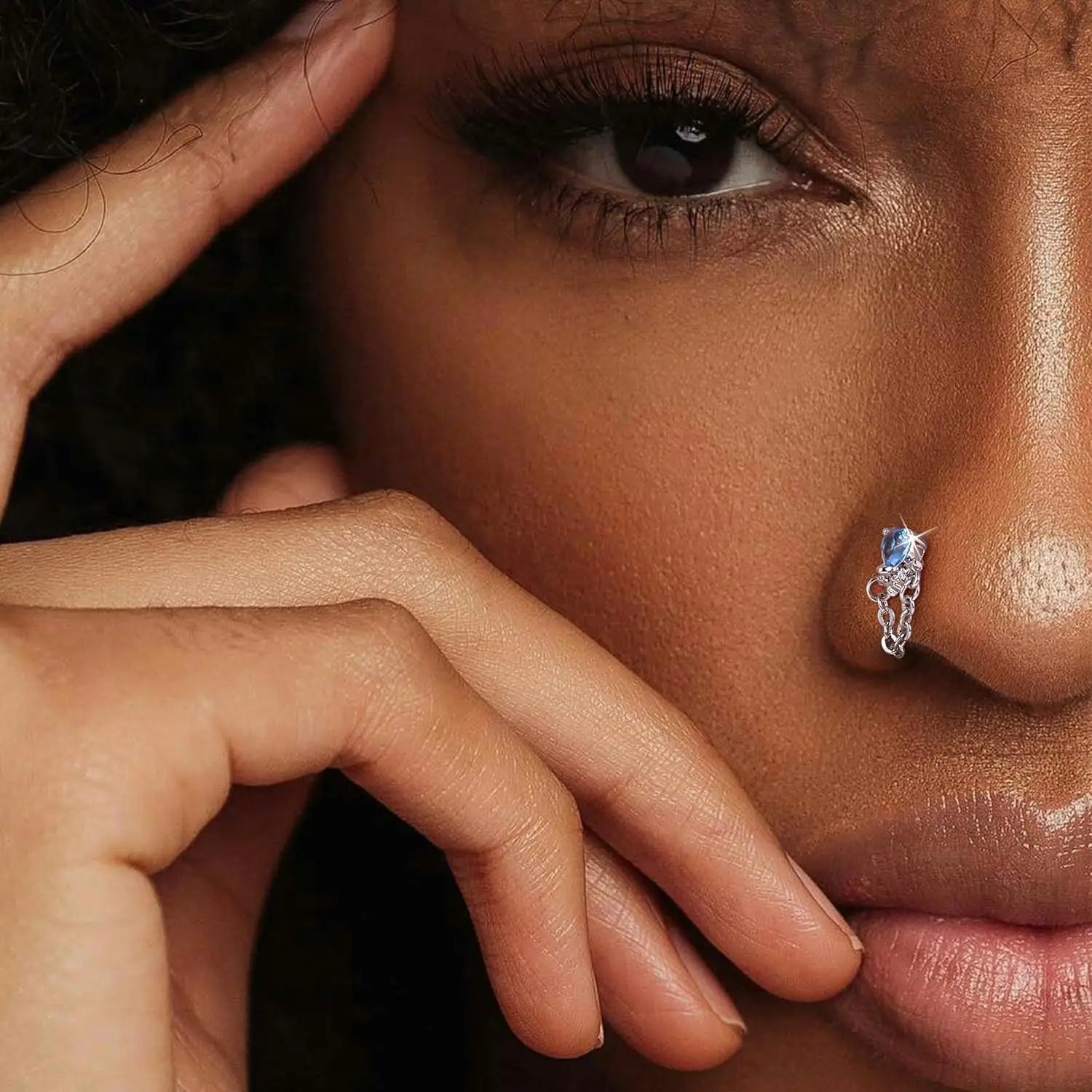 Close-up of a woman's face with a hand on her cheek, wearing a nose ring with a blue gemstone.
