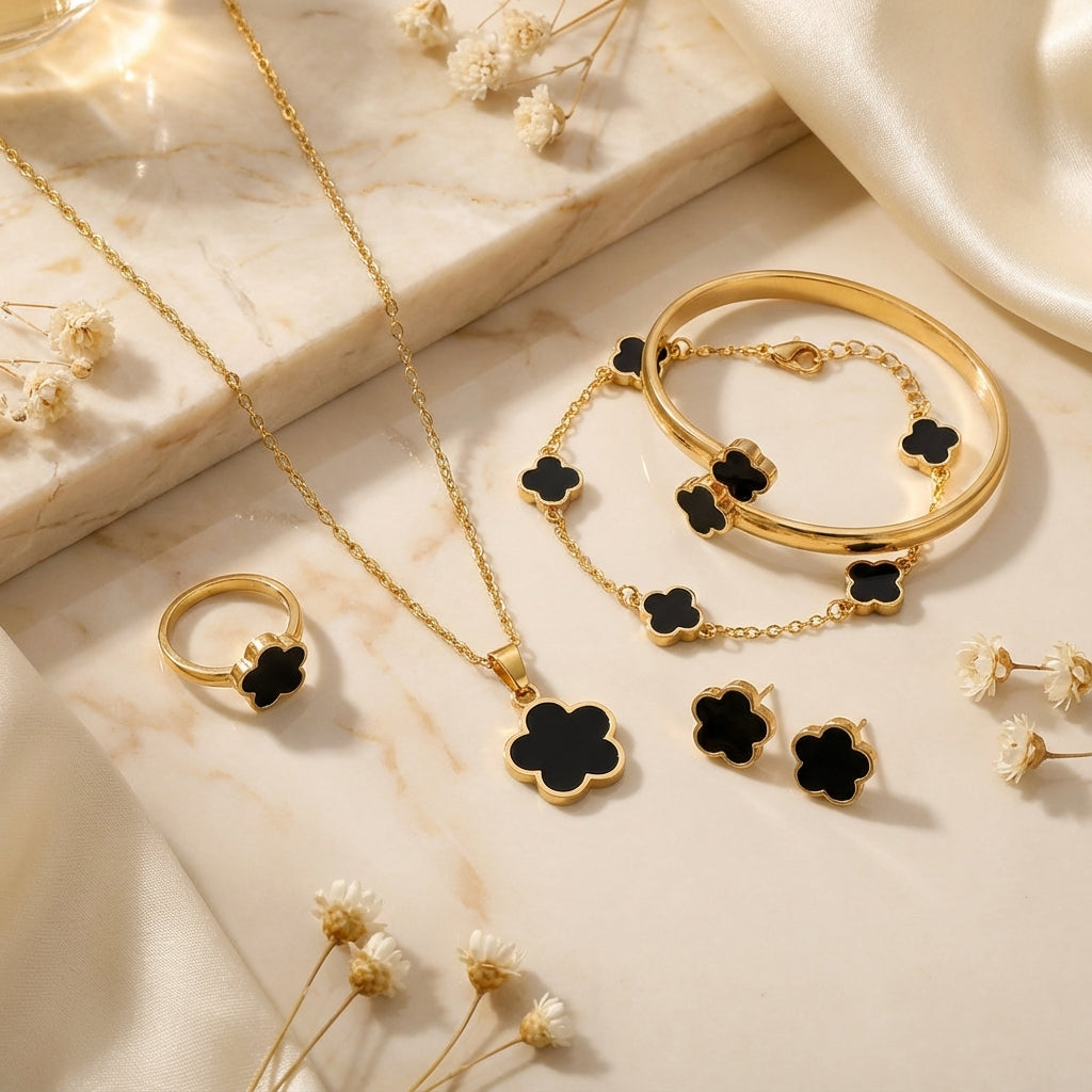Gold jewelry set with black clover accents on a marble surface with dried flowers.